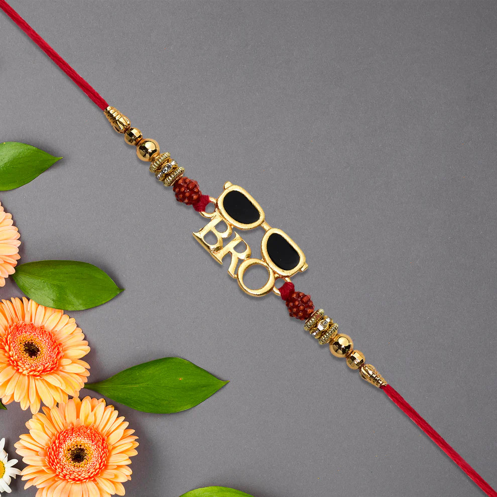 A gold-plated 'BRO' rakhi featuring a sunglasses motif with black lenses, accented with red beads and gold spacer beads on a red cord, styled with orange flowers and green leaves on a gray background.