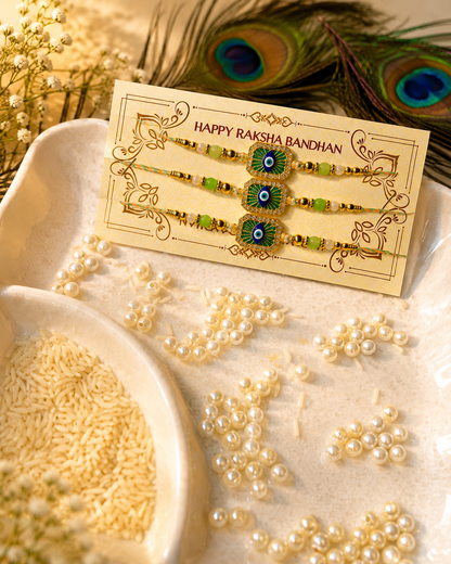 Three Rakhi bracelets displayed on a decorative cream card that reads 'HAPPY RAKSHA BANDHAN.' The card is placed on a tray with scattered pearl-like beads, rice, and peacock feather accents.
