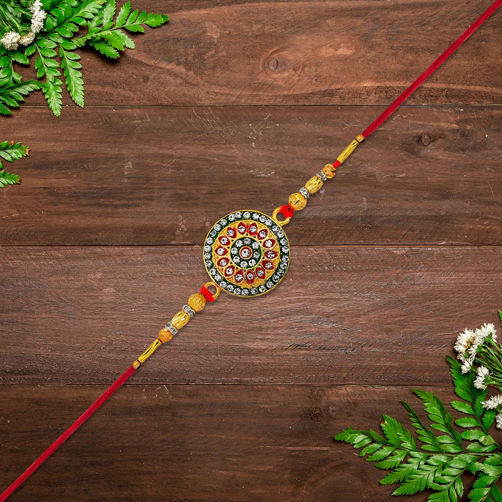 A traditional Indian Rakhi with a circular mandala charm, red strings, and gold and red beads, laid diagonally on a dark brown wooden background with decorative green fern leaves in the corners.