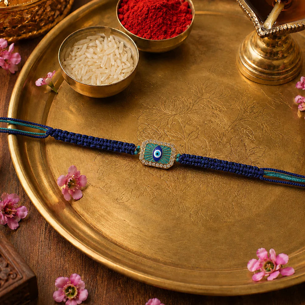 A blue braided string bracelet with a central evil eye charm, laid out on a round gold-colored tray. The tray is styled with small bowls of white grains and red powder, accented with pink flowers.